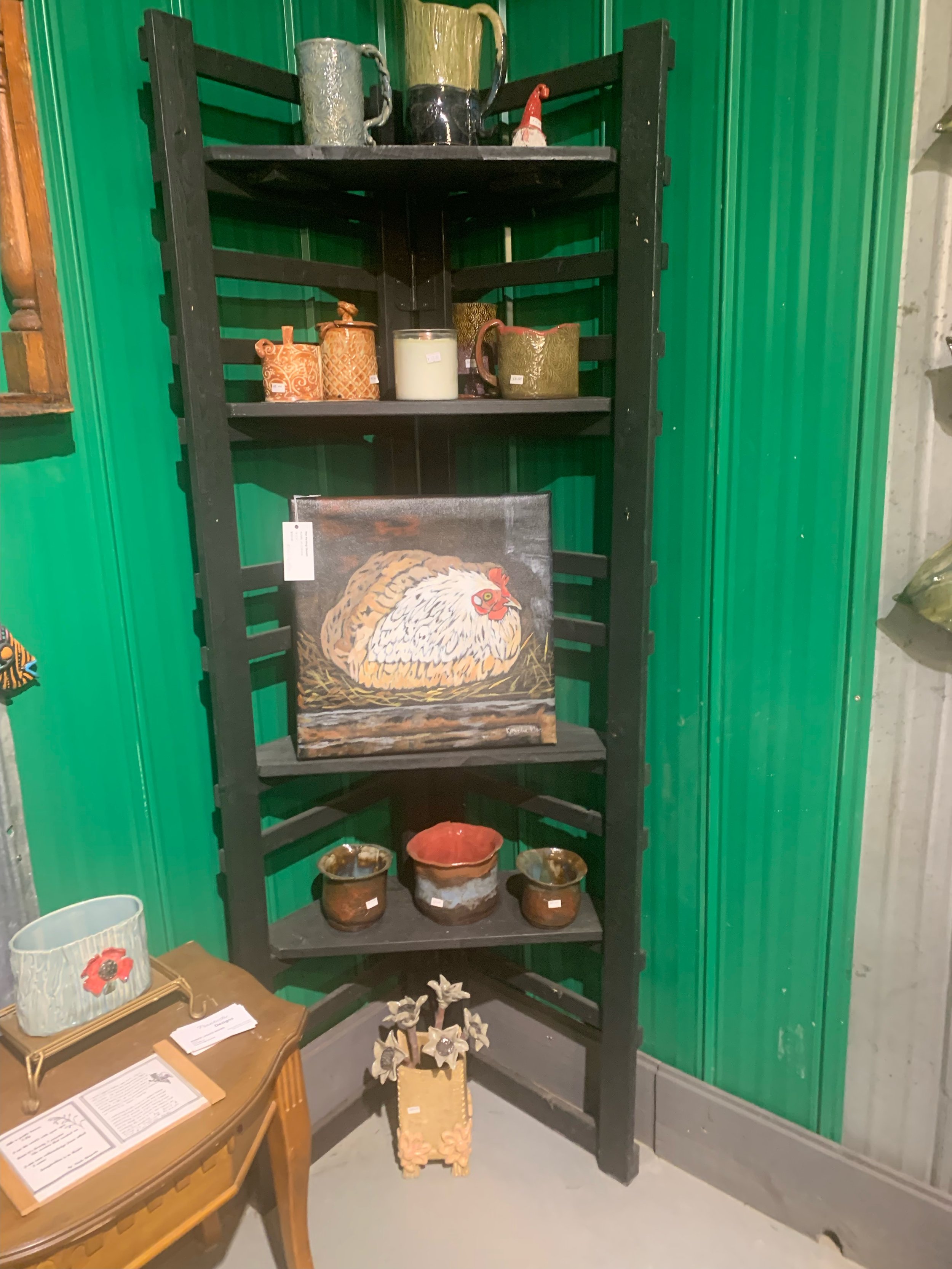 A black wooden corner shelf displaying various decorative items including ceramic mugs, bowls, a painted chicken artwork, small plants, and a large tin jug. The background shows green corrugated metal panels.