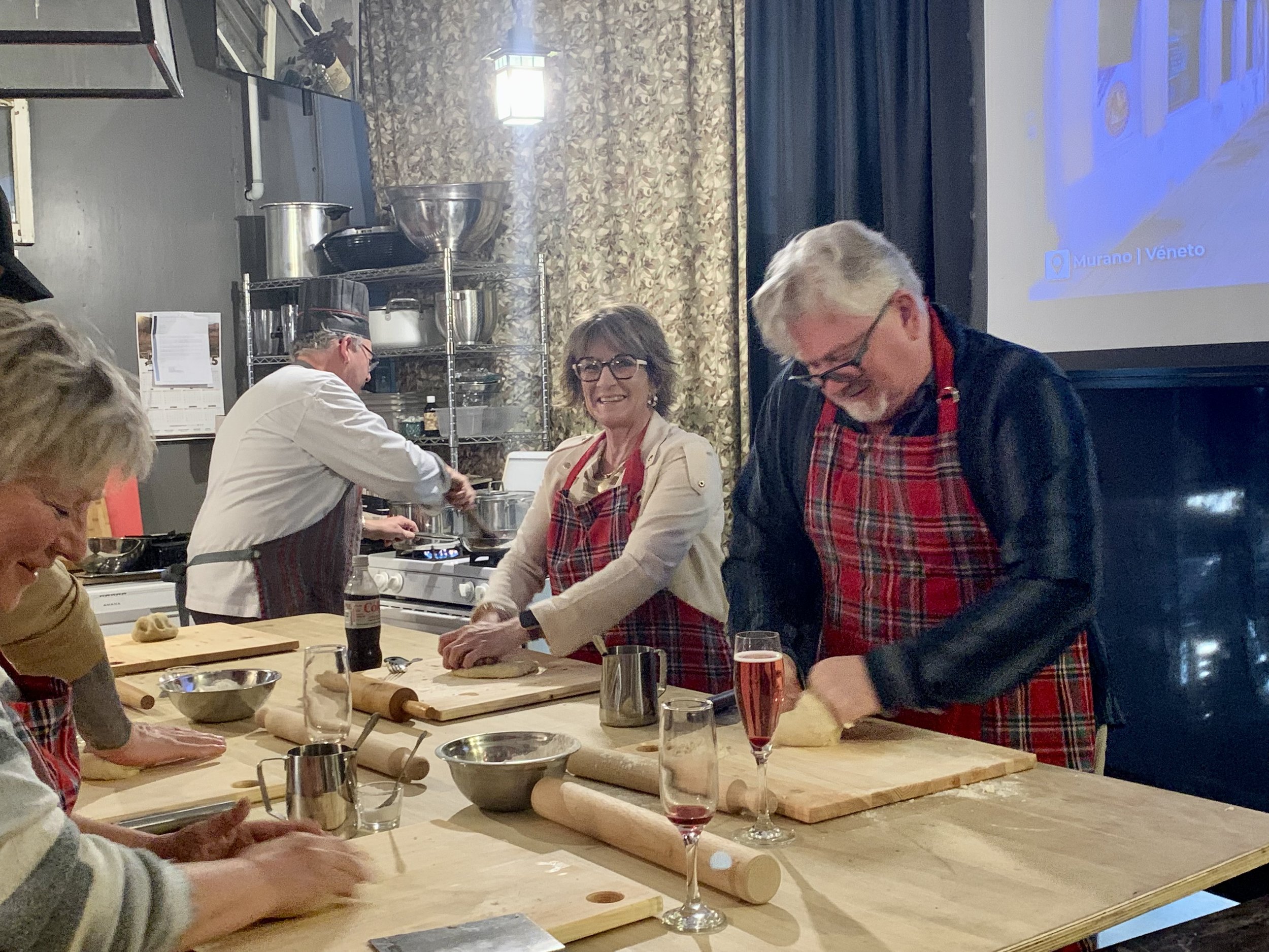 Group of people in a kitchen preparing dough, wearing aprons, with bowls and rolling pins on a wooden countertop.