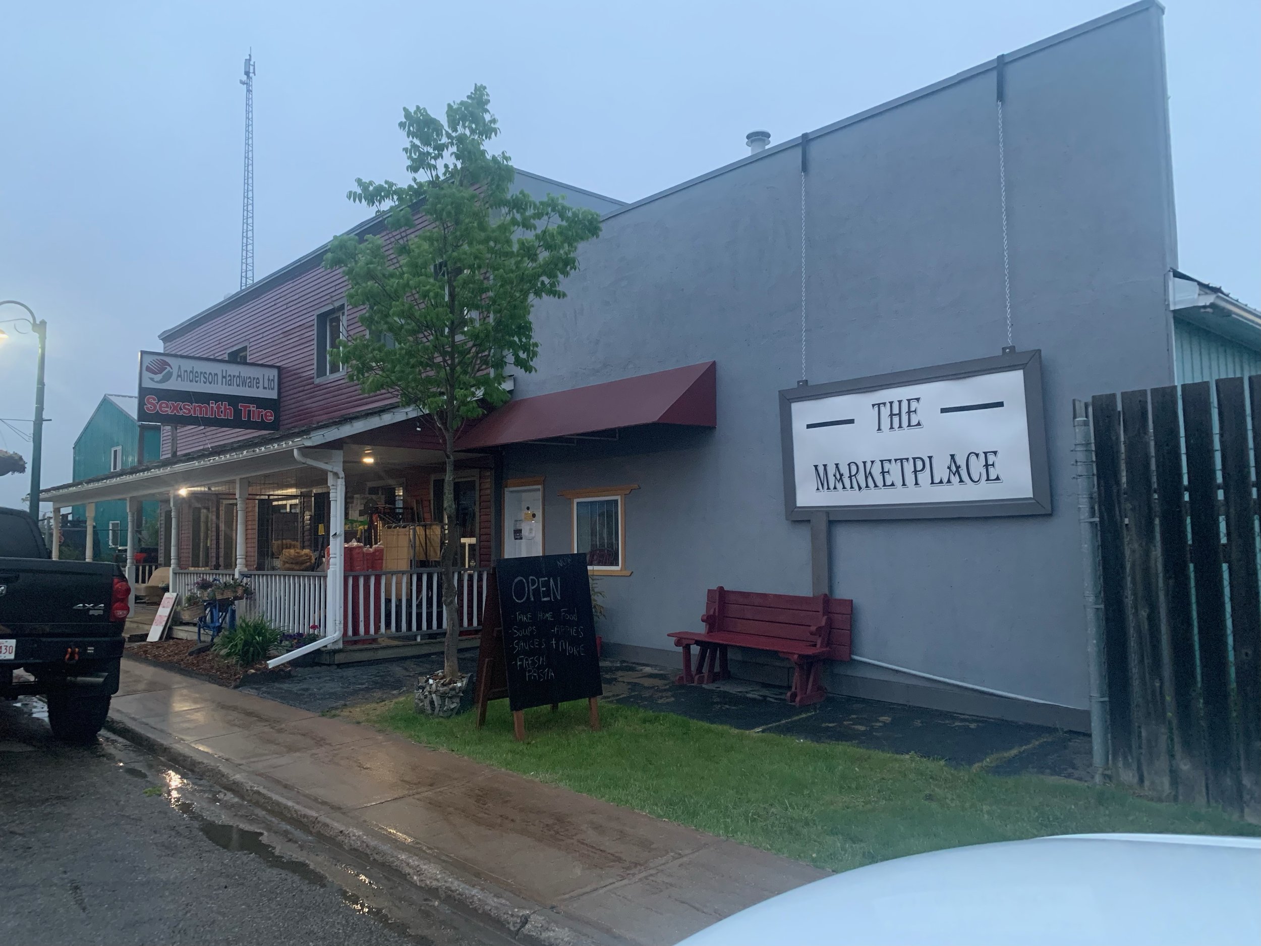A small business storefront with a sign that reads 'THE MARKETPLACE', a blackboard sign, a bench, and other signs and decorations outside, in a light rain.