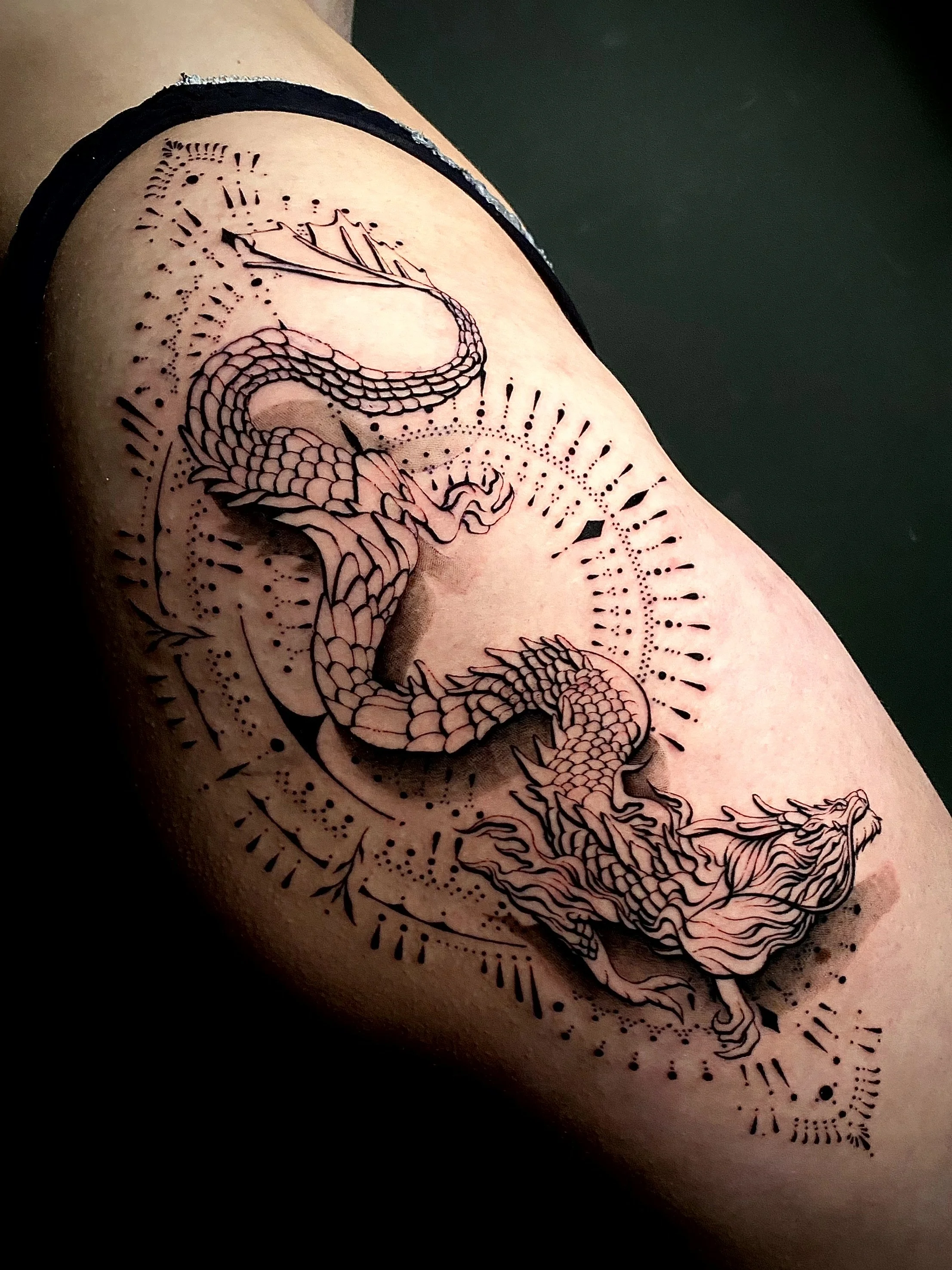 Tatouage d'un dragon stylisé en noir et gris sur le bras d'une personne, entouré de motifs géométriques et de points.