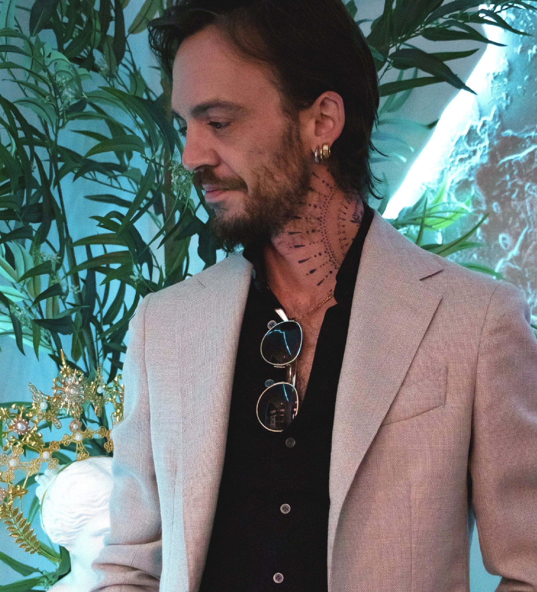 Un homme avec des cheveux longs, une barbe et des boucles d'oreilles, portant un blazer clair et une chemise noire, se tient à côté d'une décoration dorée et d'une plante verte, dans un environnement avec un éclairage bleu et vert.