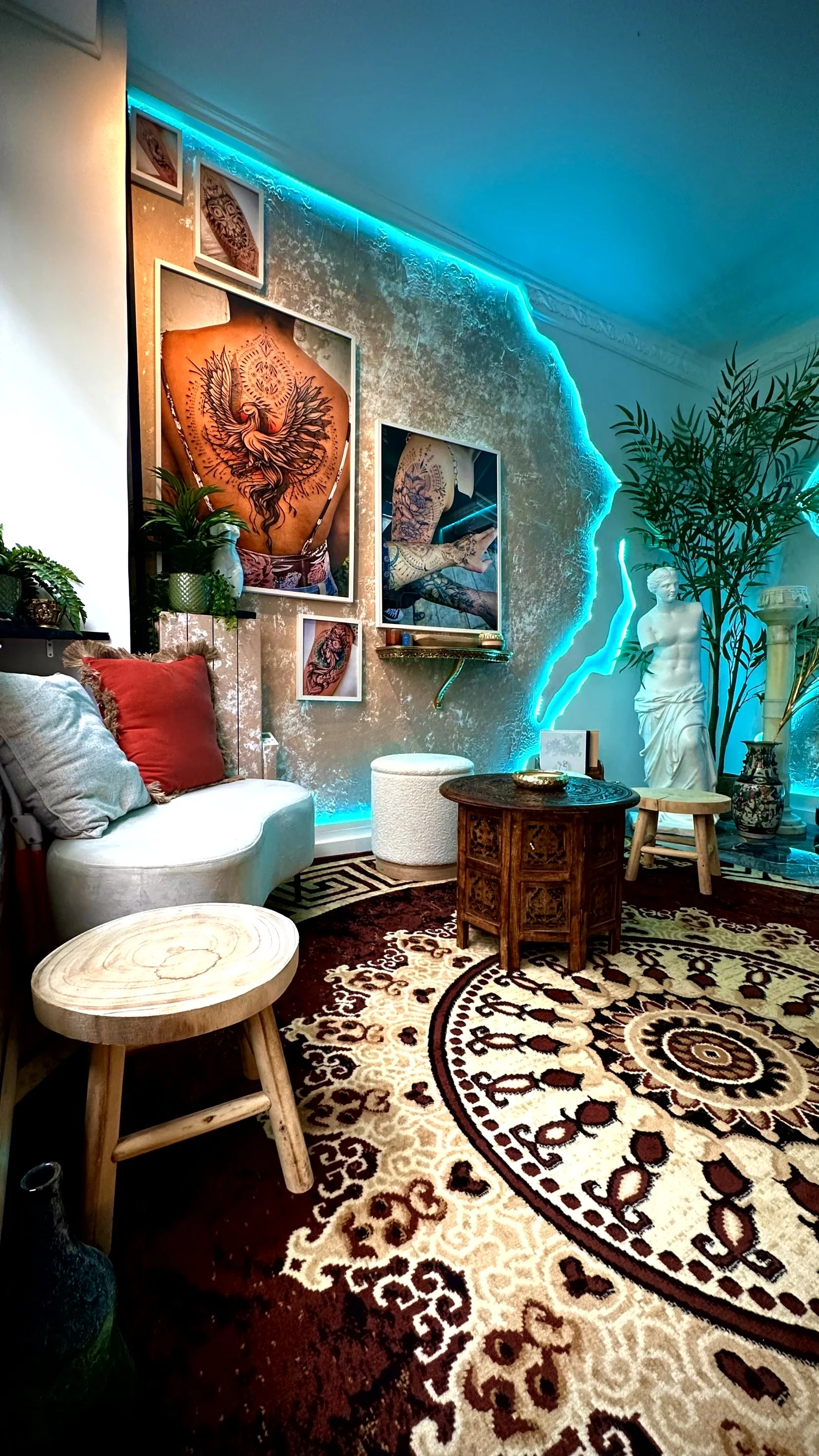 Salon éclectique avec mobilier vintage, tapis ornemental, œuvres d'art tattoo, statues, plantes et éclairage LED bleu.