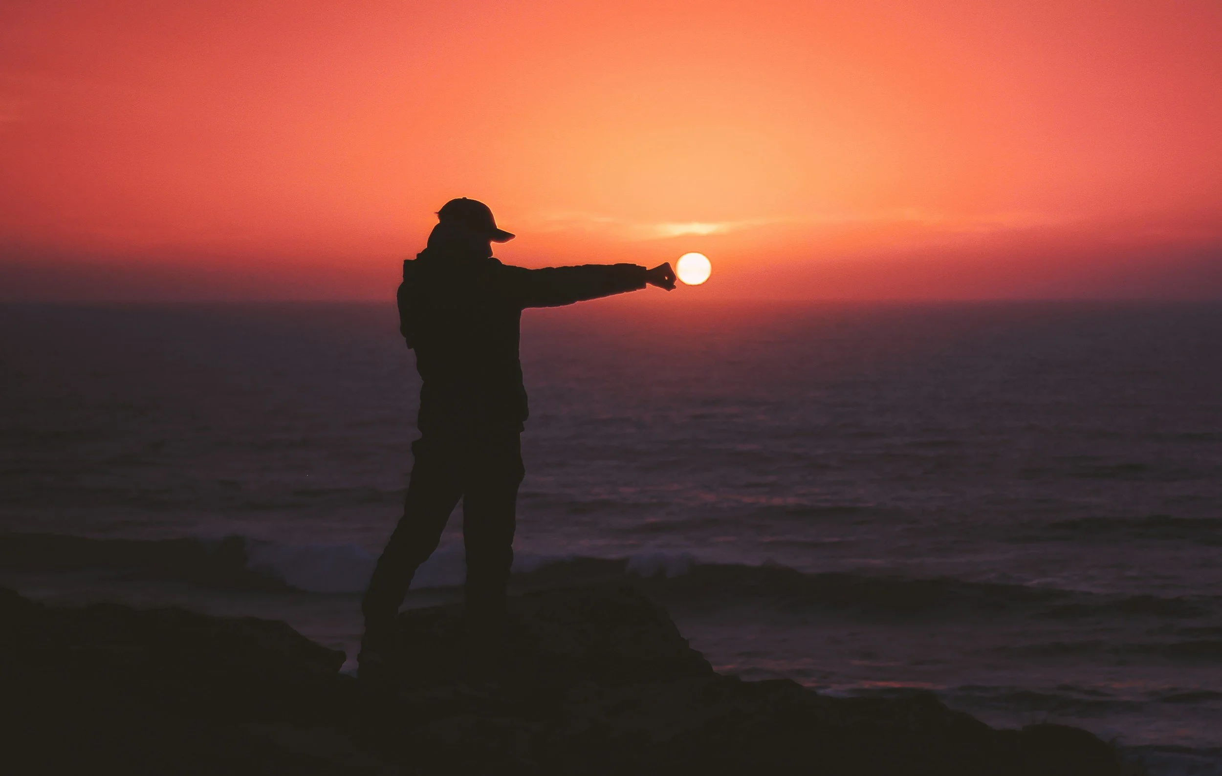 Silhouette of a person wearing a hat standing on rocks by the ocean during sunset, holding the sun between their hand and arm.