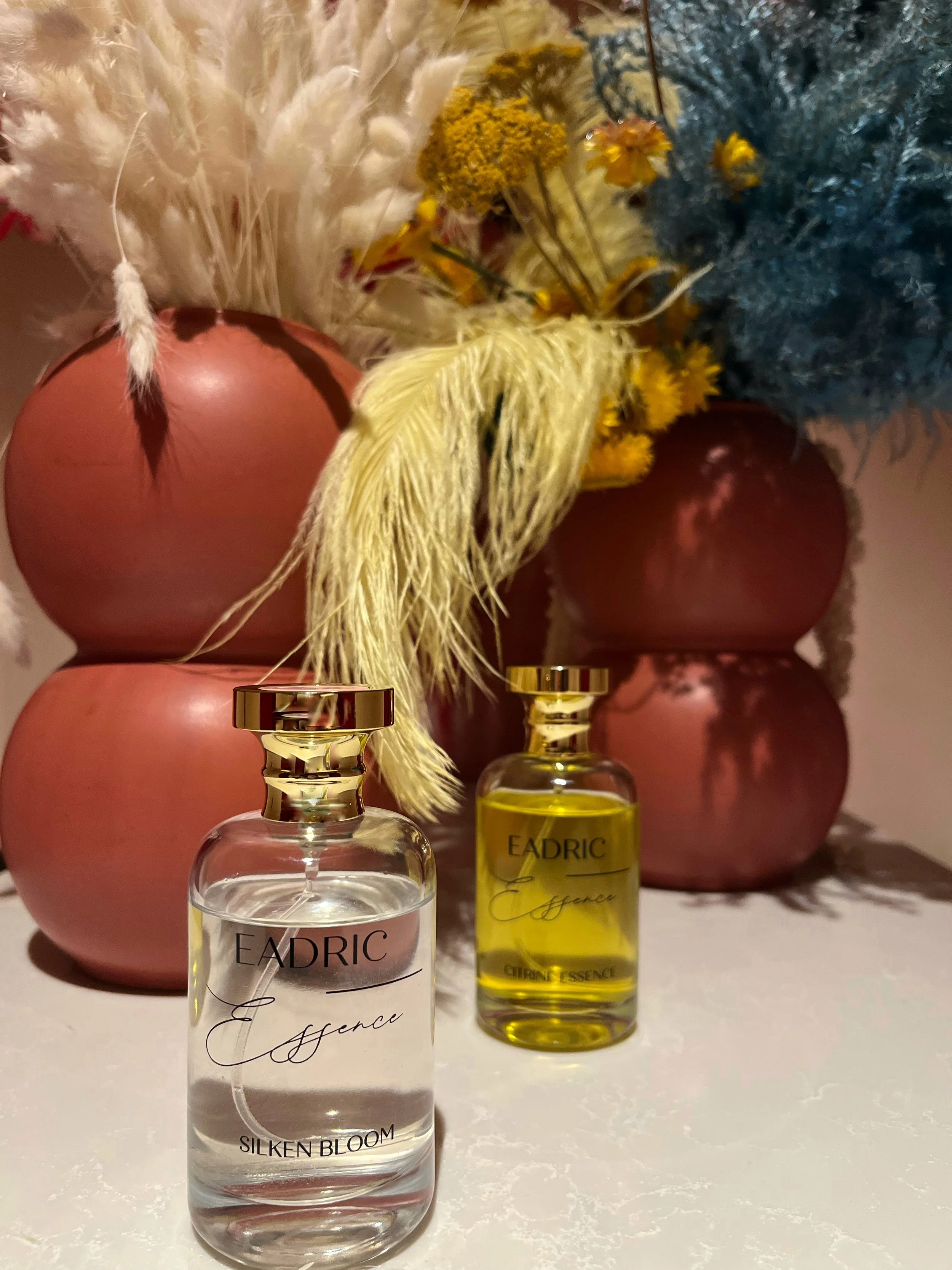 Two bottles of Eadric Essence perfume, one with clear liquid labeled 'Silken Bloom' and the other with yellow liquid, placed on a white surface in front of pink vases filled with dried flowers and plants, including yellow, cream, and blue foliage.