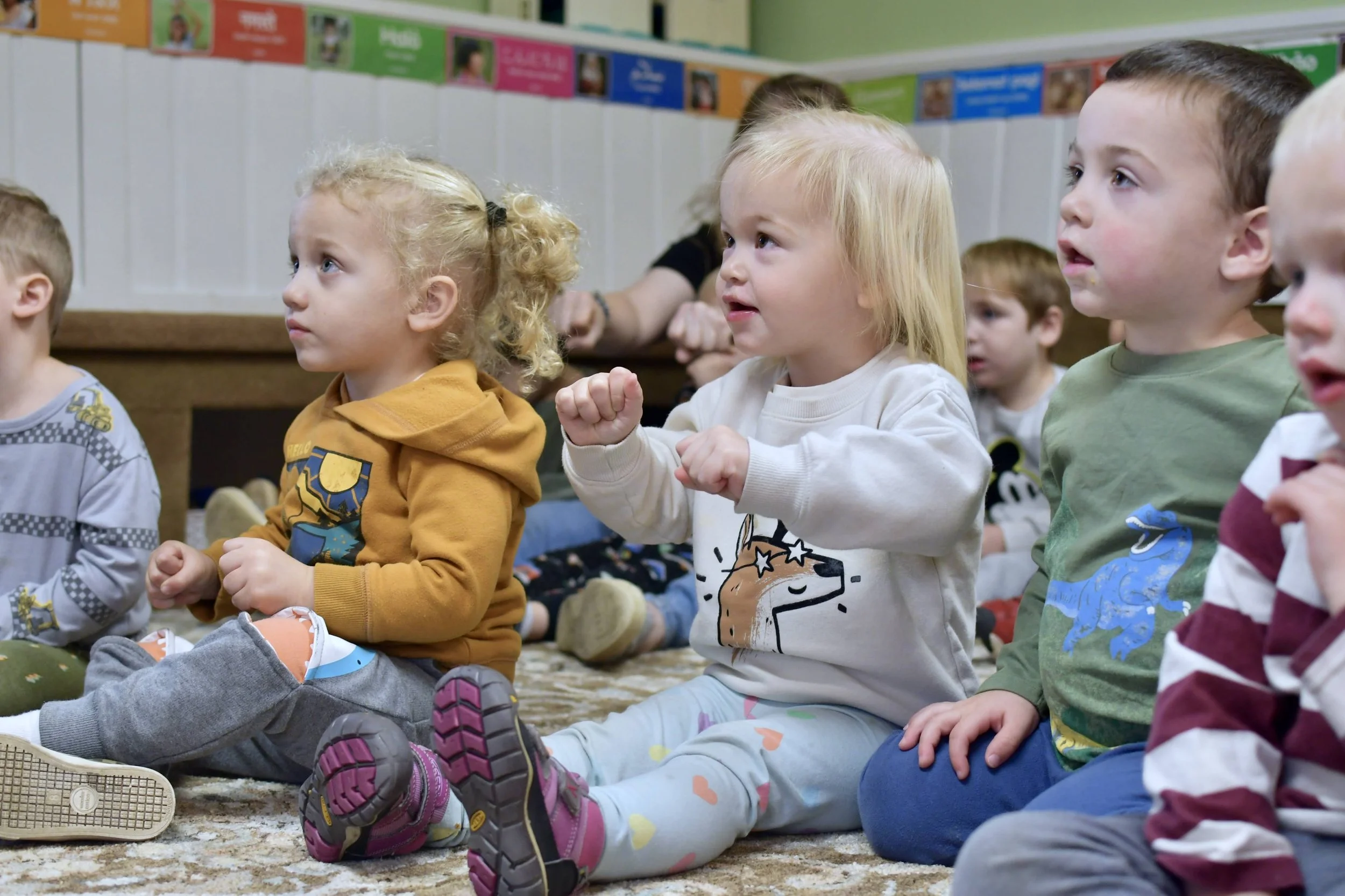 Group of young children sitting on the carpeted floor in a classroom, attentively watching a presentation or performance, with some children making excited gestures.