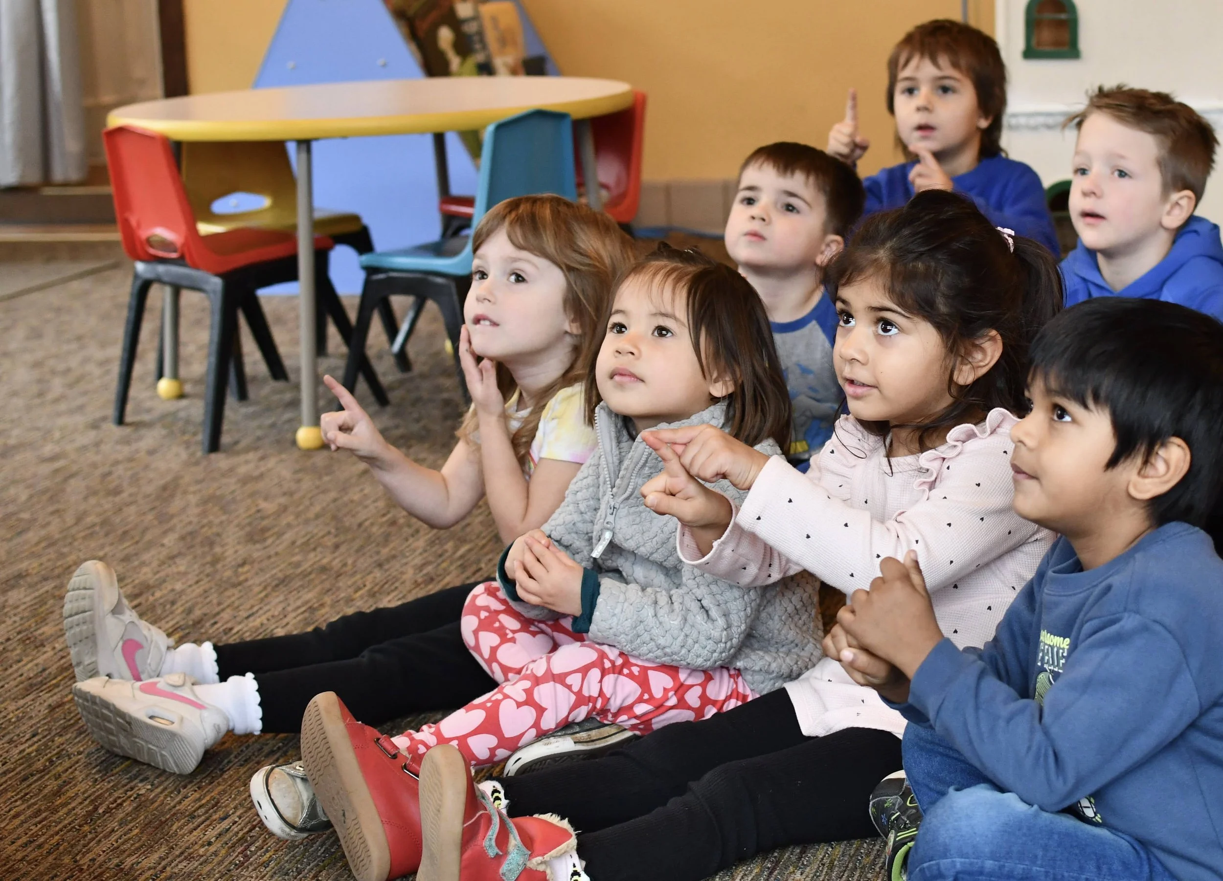 Group of young children sitting on the floor in a classroom, watching and listening attentively to something out of the frame.