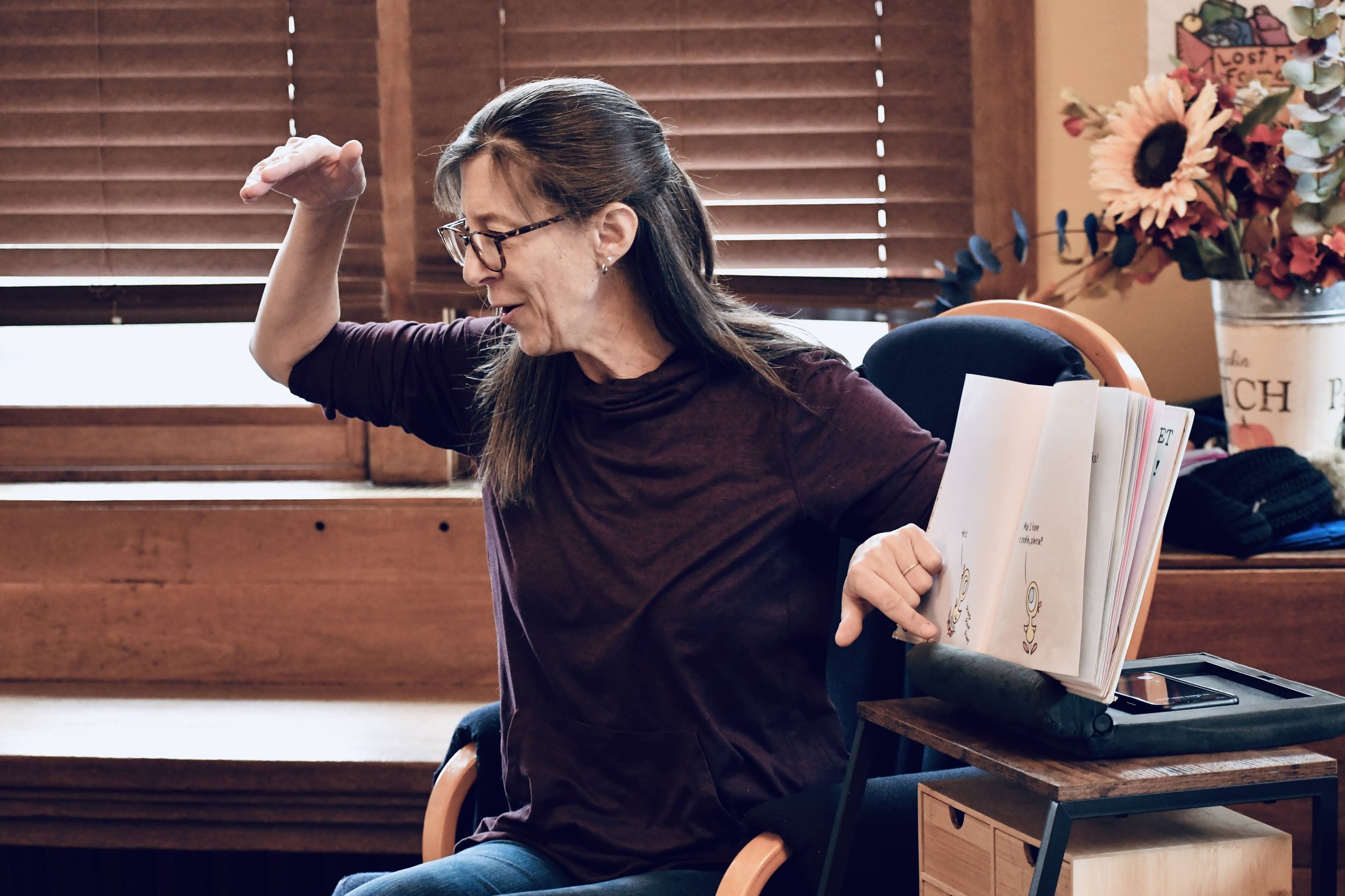 A woman with glasses and long dark hair sits on a chair, appearing to stretch or exercise, with a smile on her face. She is in a room with wooden blinds, a small wooden table holding a laptop and a stack of books or notebooks, and a large flower arra