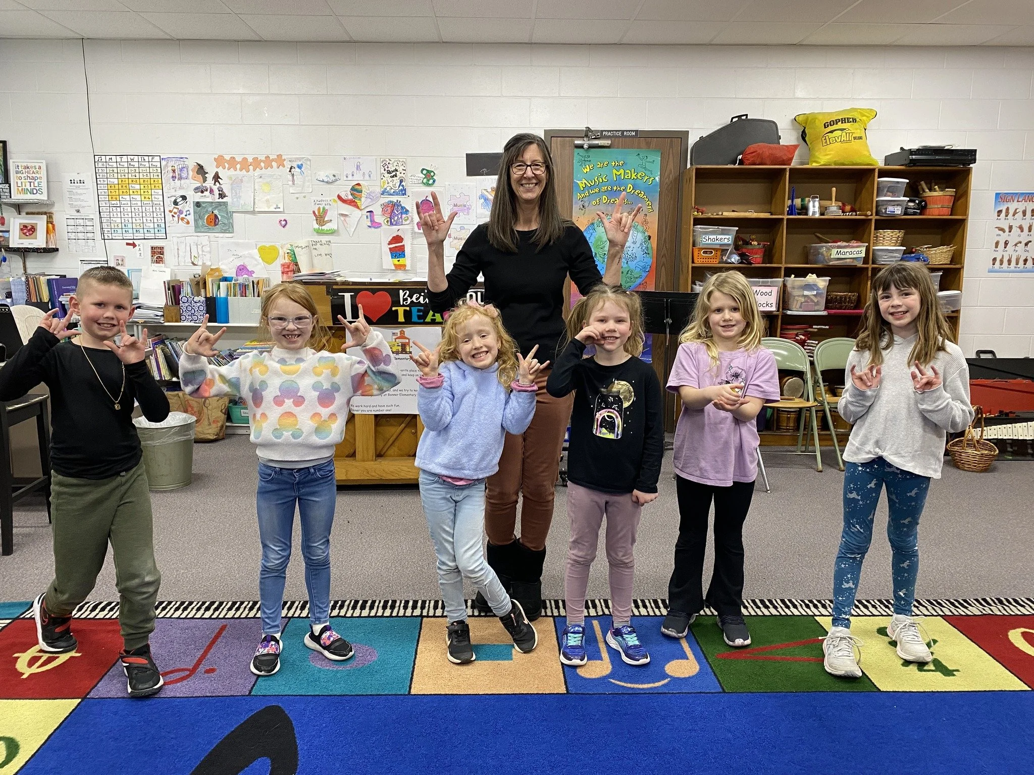 A group of seven children and one adult in a classroom, smiling and making peace signs, standing on a colorful alphabet rug.