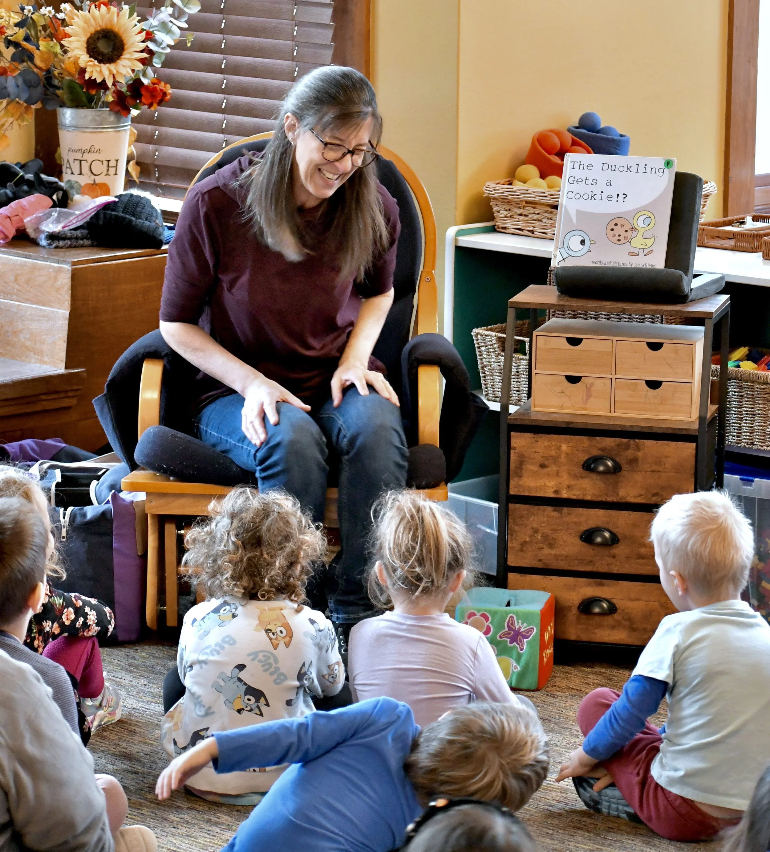 A woman with glasses is sitting on a chair, engaging with a group of young children sitting on the floor, during a storytime or educational activity in a cozy, well-lit room with wooden decor, colorful baskets, and a sign that reads 'The Duckling Get