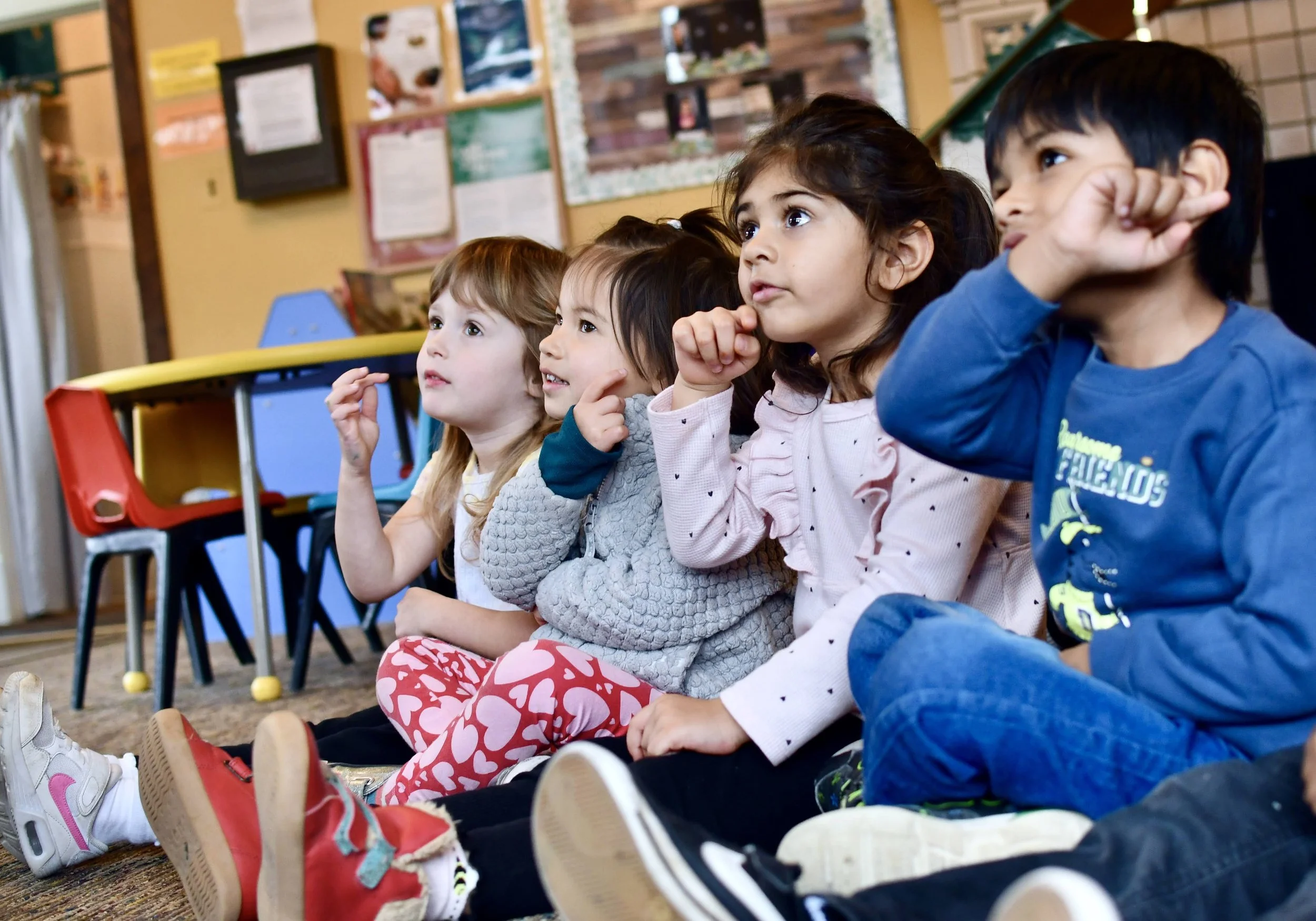 Five young children sitting on the floor in a classroom, attentively watching something with interest.