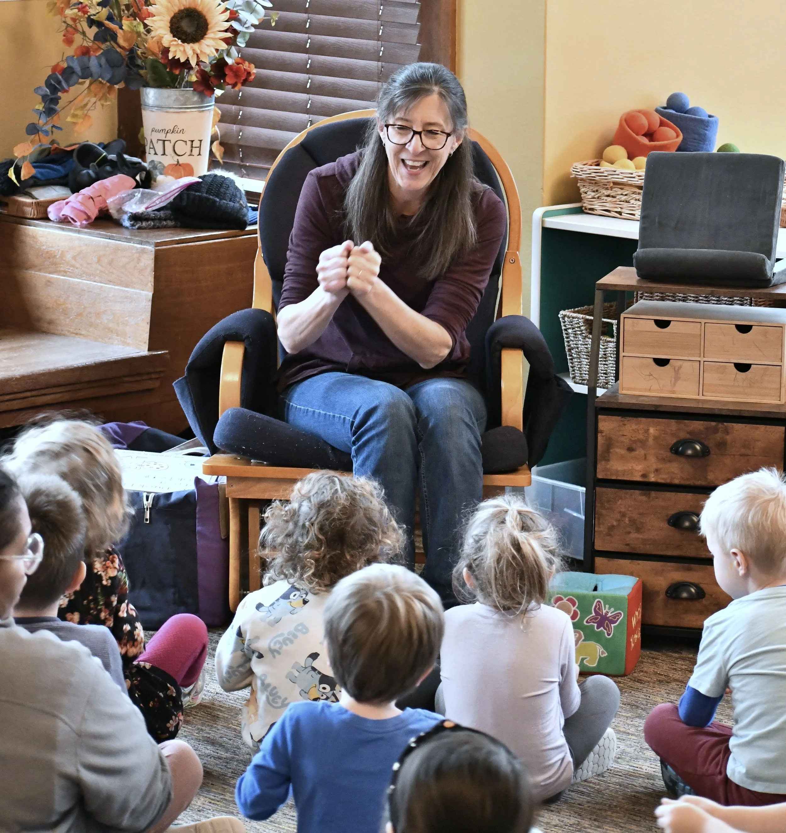 A woman with glasses sitting in a chair, engaging with a group of children sitting on the floor in front of her, in a cozy indoor setting with wooden furniture and colorful decorations.