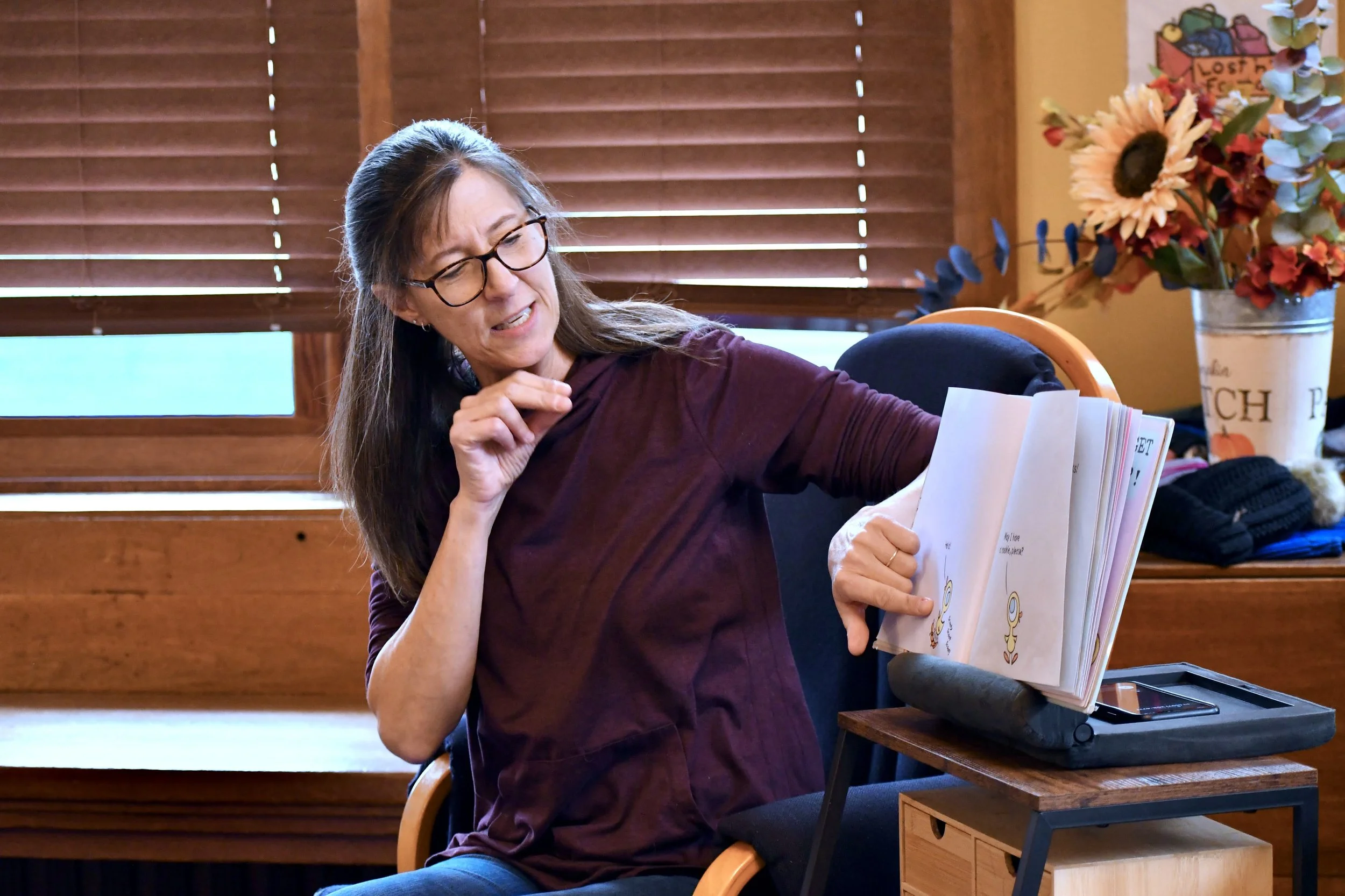 A woman with glasses and long brown hair is sitting in a wooden-framed chair, turning the pages of a book that is placed on a cushion on her lap. She appears to be mid-conversation or explaining something. Behind her, there are wooden window blinds a