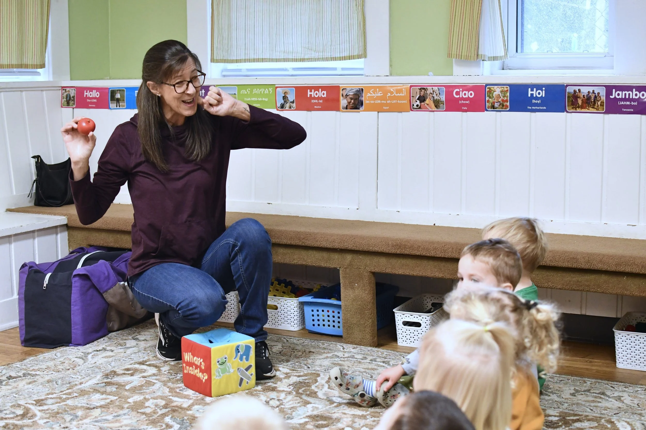 A woman with glasses and long brown hair, wearing a maroon shirt, kneeling on a carpeted floor, holding a red apple, teaching a group of young children in a classroom. The children are sitting on the carpet, facing the woman, with some having blonde 