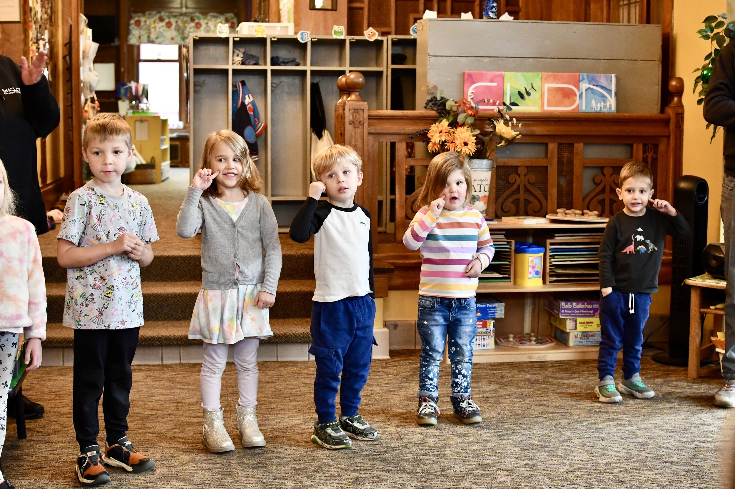 Group of young children standing in a line indoors, participating in an activity. The background features shelves with toys, books, and decor.