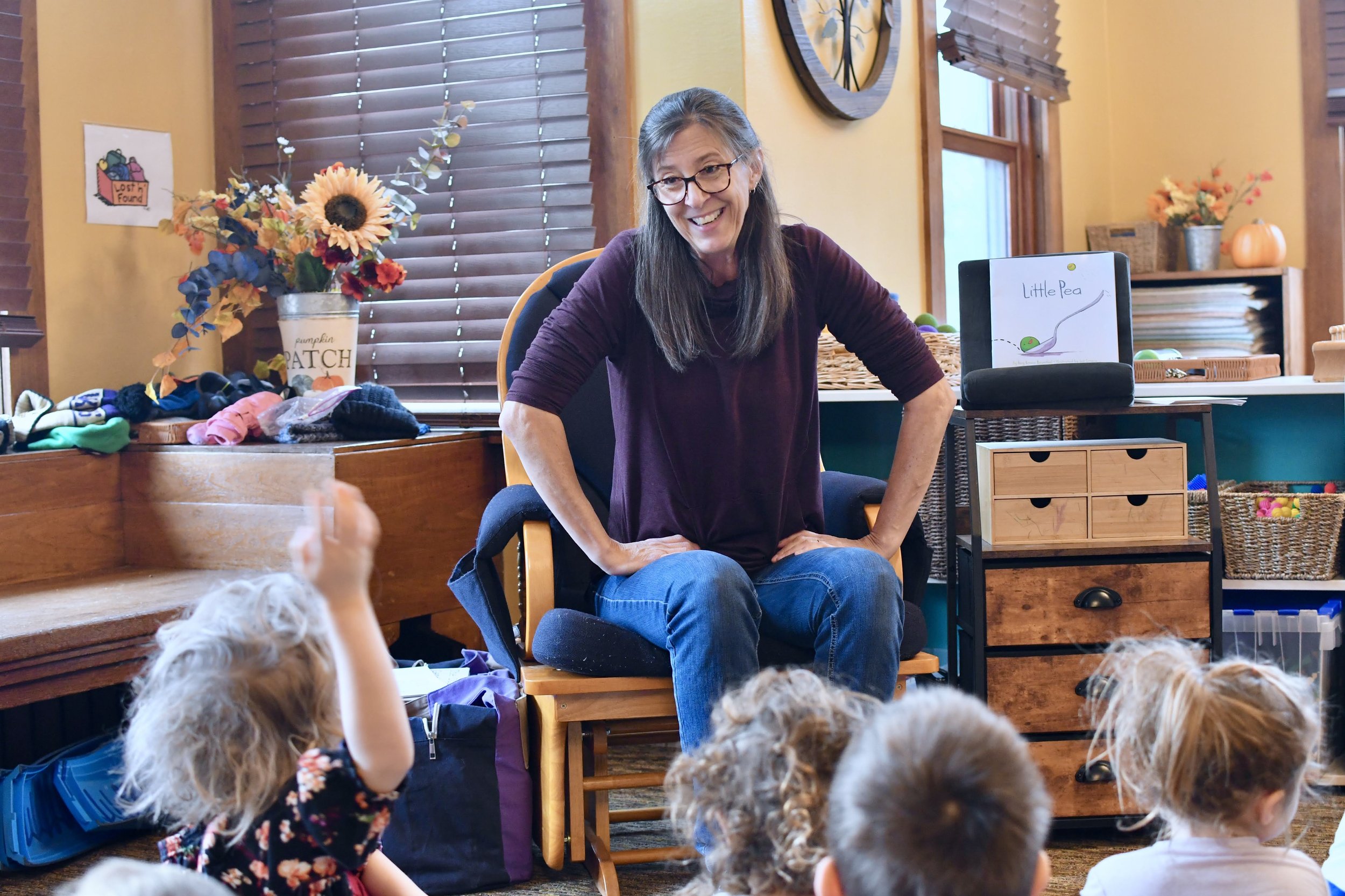 A woman with long dark hair and glasses sitting on a wooden chair, smiling and engaging with a group of young children in a cozy classroom setting decorated with flowers and educational materials.