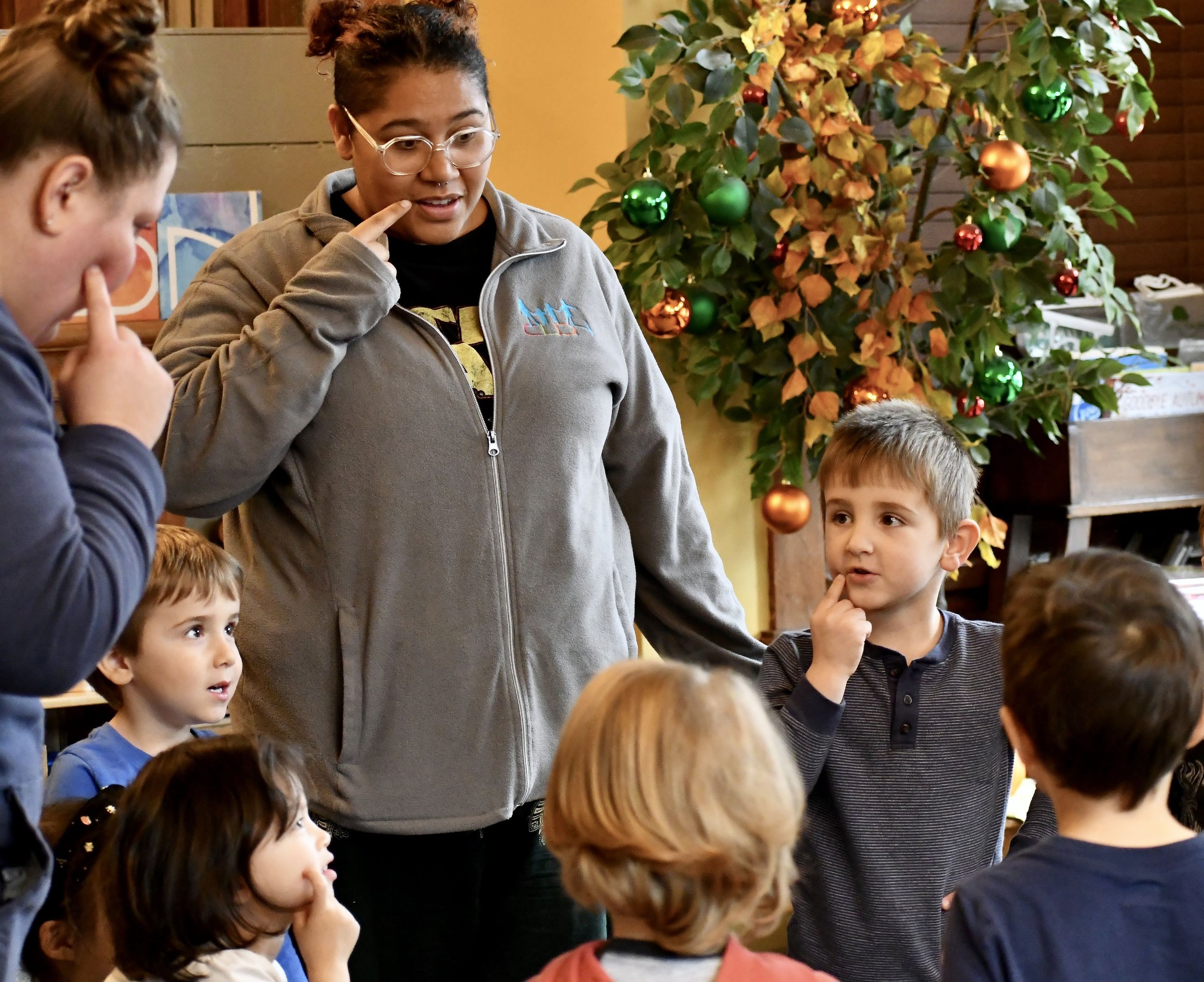 A group of children and an adult woman with glasses and a gray zip-up jacket are indoors, in front of a decorated Christmas tree, engaging in a conversation or activity.