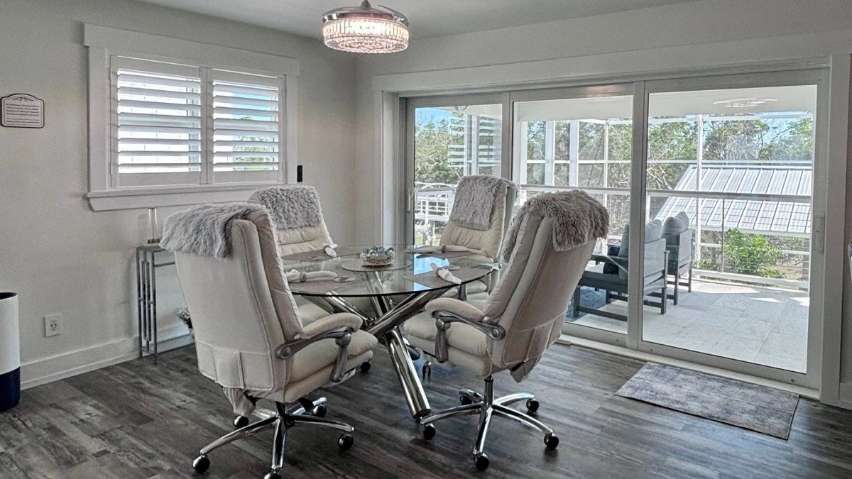 Dining room with four white chairs around a glass table, large sliding glass door opening to an outdoor deck, and a chandelier overhead.