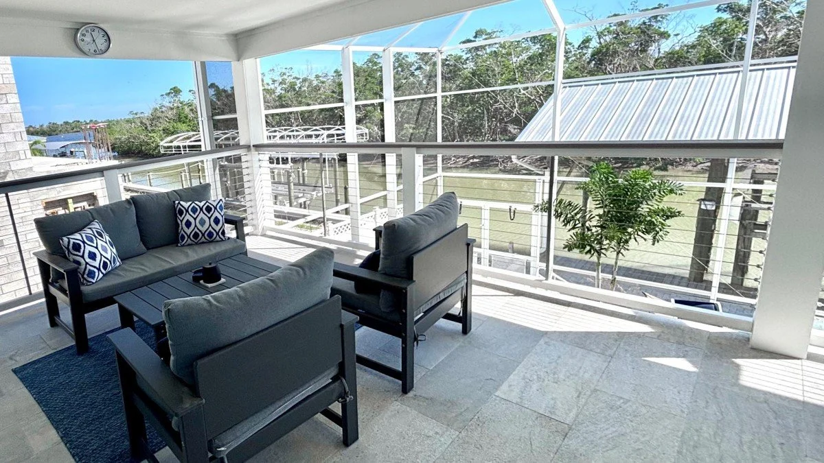 A screened porch with a seating area featuring outdoor furniture, a clock on the wall, and a view of trees and a waterway outside.