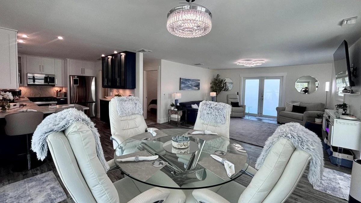 Open-concept living and dining room with a glass dining table and white chairs, a kitchen with stainless steel appliances, and a living area with sofas, a TV, and sliding glass doors.