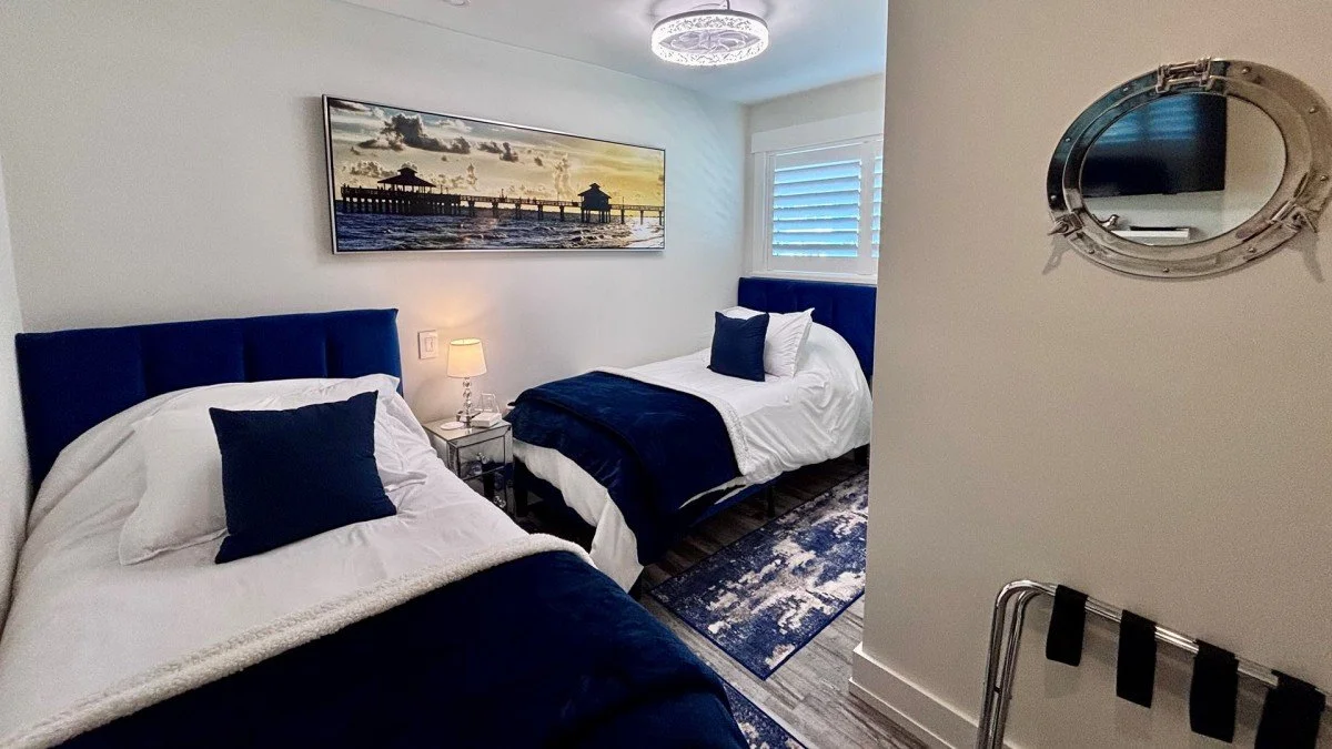 A bedroom with two twin beds, each with white bedding and navy blue pillows and headboards. There is a small nightstand with a lamp between the beds. A framed picture of a pier at sunset hangs on the wall. A window with blue blinds is beside one bed.