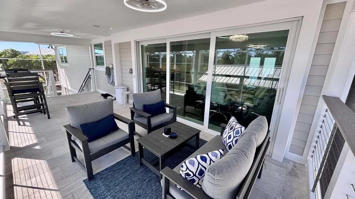 Outdoor balcony with chairs, a table, and sliding glass doors, overlooking a neighborhood with trees and rooftops.