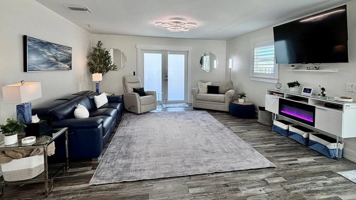 Living room with a black leather sofa, two beige armchairs, a large area rug, wall art, a wall-mounted TV, and decorative plants.
