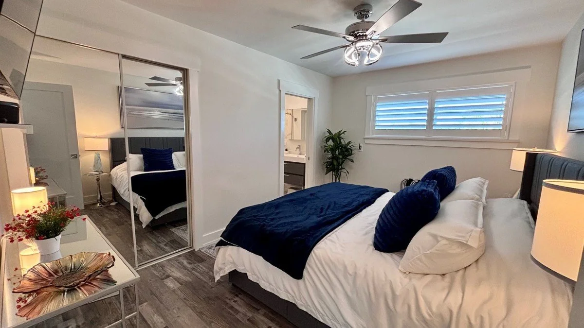 A bedroom with a large bed covered in white and navy blue bedding, two navy blue pillows, a window with shutters, a ceiling fan, a potted plant, and a mirrored closet door reflecting part of the room.