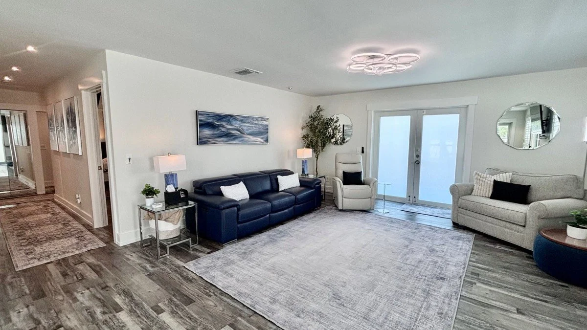 Living room with white walls, dark blue leather sofa, white armchair, light gray sofa, large area rug, modern ceiling light fixture, two side tables with lamps, round mirror, potted plants, sliding glass doors, and hardwood floors.