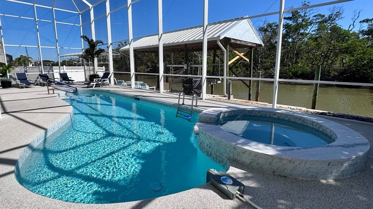 A screened-in backyard pool and hot tub with outdoor furniture and a view of a river in the background under a clear blue sky.