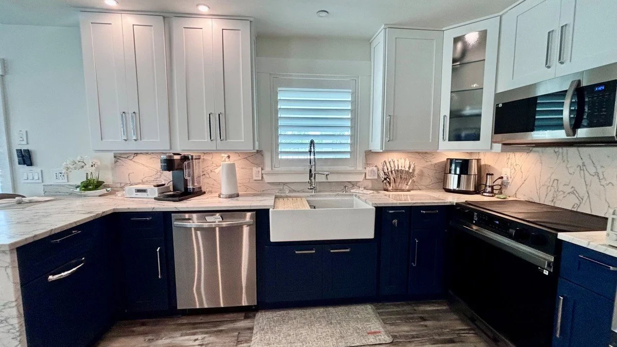 Modern kitchen with white upper cabinets, navy lower cabinets, marble countertops, and a farmhouse sink. Appliances include a dishwasher, a microwave, a coffee maker, a toaster, and an oven. There is a window with blinds above the sink and decorative