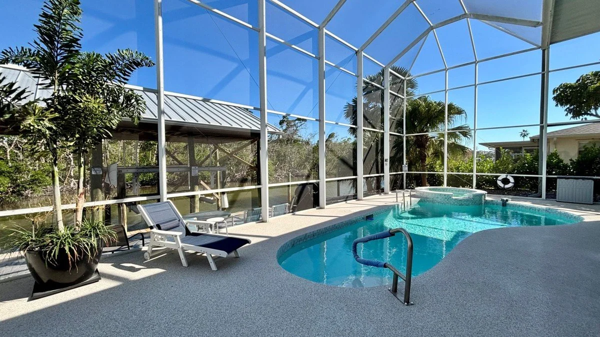 Indoor pool area with lounge chairs, potted plant, and glass enclosure showing blue sky and palm trees outside.