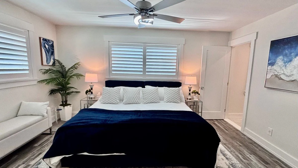 A bedroom with a bed, white bedding, and multiple pillows. Nightstands with lamps on each side of the bed. A window with white shutters above the bed. A ceiling fan overhead. A white sofa with a pillow on the left, a large plant near it, and artwork 