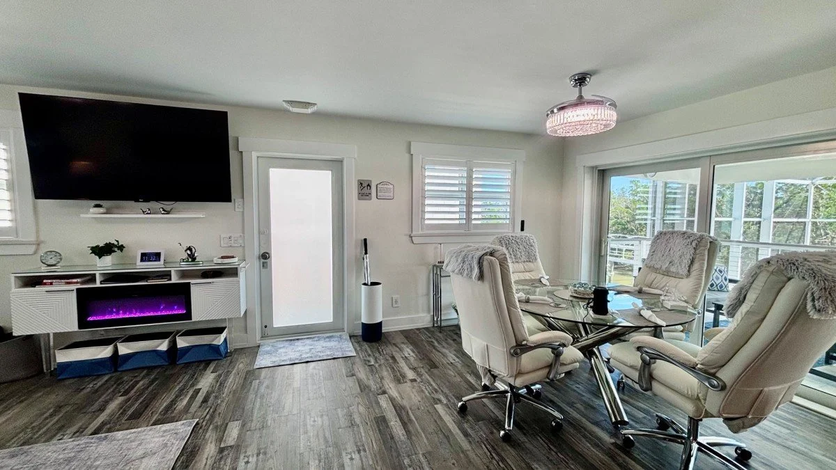 A living room with a mounted flat-screen TV, a white entertainment console with a glowing purple electric fireplace, a glass dining table surrounded by padded beige chairs, a sliding glass door leading to a balcony, and windows with white shutters. T