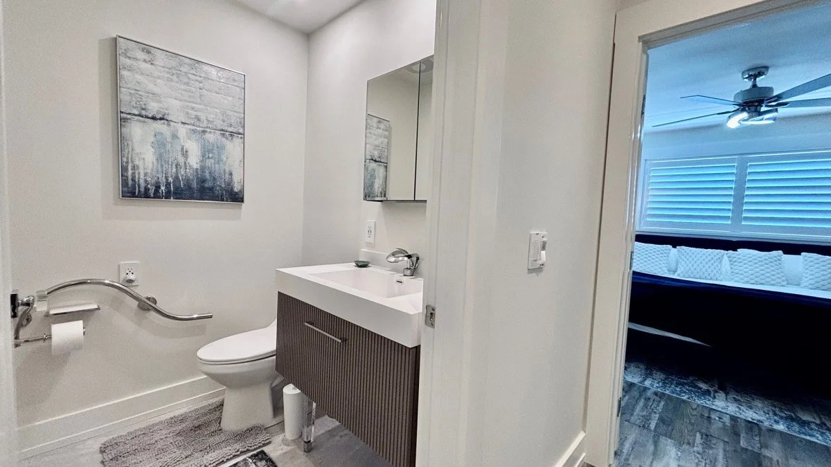 A small bathroom with a modern sink vanity, a mirror, and a toilet. There is a piece of artwork on the wall and a textured rug on the floor. The bathroom door is open, revealing a bedroom with a ceiling fan and white window shutters.