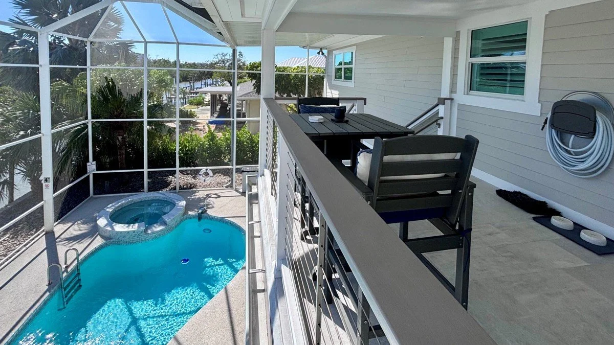 View of a screened-in patio with a small, curvy swimming pool and a hot tub, overlooking trees and a body of water beyond, with outdoor furniture and a coiled garden hose on the wall.