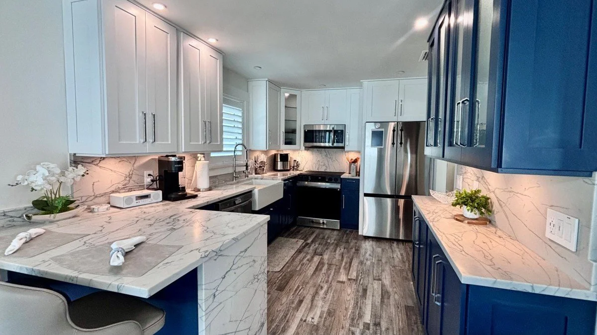 Modern kitchen with white upper cabinets and navy lower cabinets, marble countertops, stainless steel appliances, hardwood flooring, and a nearby dining area with place settings.