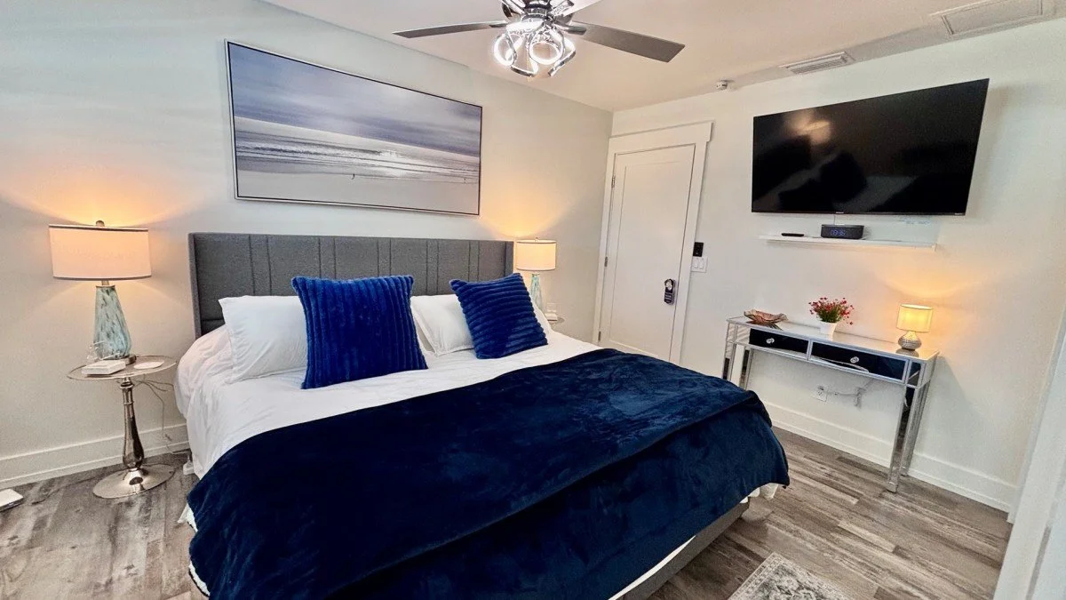 A bedroom with a bed, blue pillows, white sheets, and a dark blue blanket. Two bedside lamps on glass tables, wall art above the bed, a wall-mounted TV, and a mirrored console with a plant and decorative items.