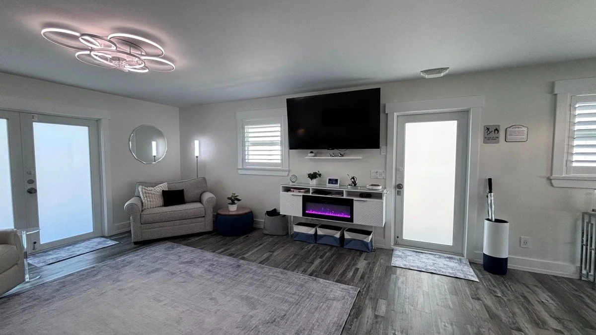Modern living room with white walls, a large flat-screen TV, a mounted electric fireplace, gray couch, ottoman, and minimalist decor, including a circular mirror, wall sconces, and contemporary lighting fixture.