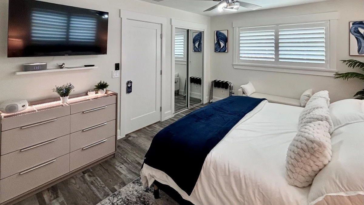 A bedroom with a large bed covered with white sheets, a navy blue blanket, and textured pillows. There's a dresser with decorative items and a wall-mounted TV. The room has windows with white blinds, framed artwork, and a mirrored closet door.
