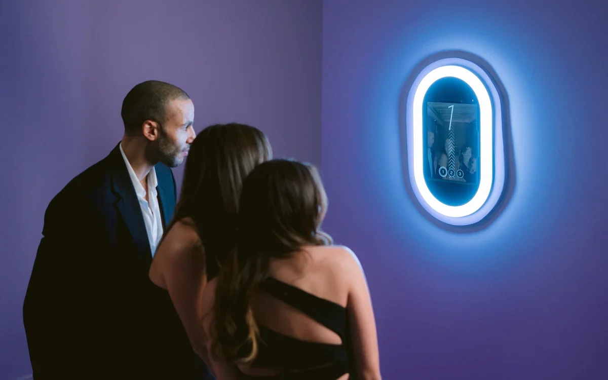 A man and two women looking at an illuminated, futuristic wall mounted photobooth in a dimly lit room.