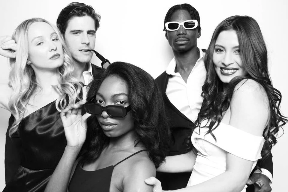 Group of five young adults posing together in stylish outfits, some wearing sunglasses, in black and white.