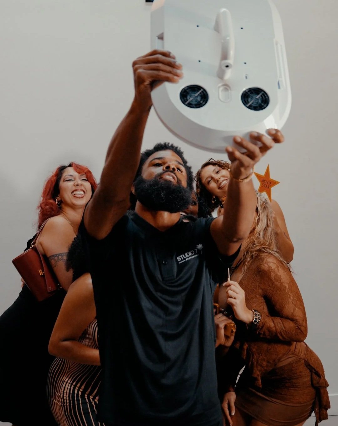 Group of people taking a selfie together, smiling and posing closely, with a plain wall background.