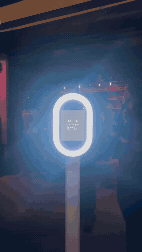 The Classic Photobooth by Studio 7 live at an event. The booth is illuminated through its ring lights with a screen saying 'tap to start'.