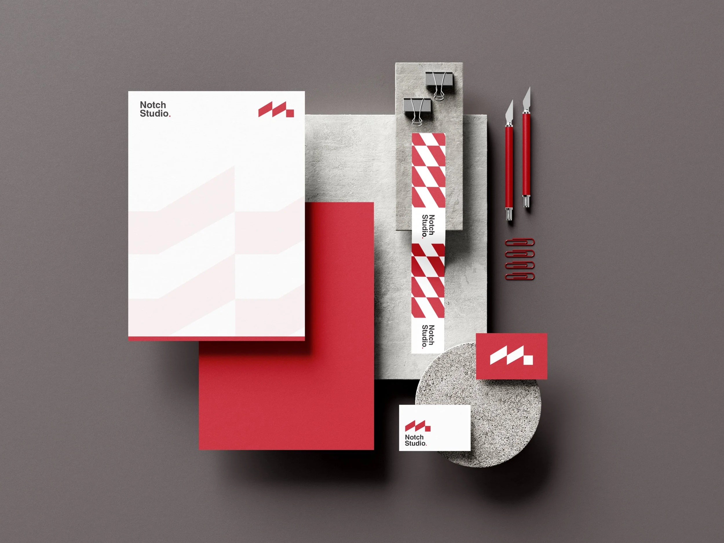 Branding materials for Notch Studio, including a white booklet, a red booklet, a white and red striped bookmark, business cards, two red pens, paper clips, and a small white card with a red and white logo, all arranged on a gray background with concrete and circular textured elements.