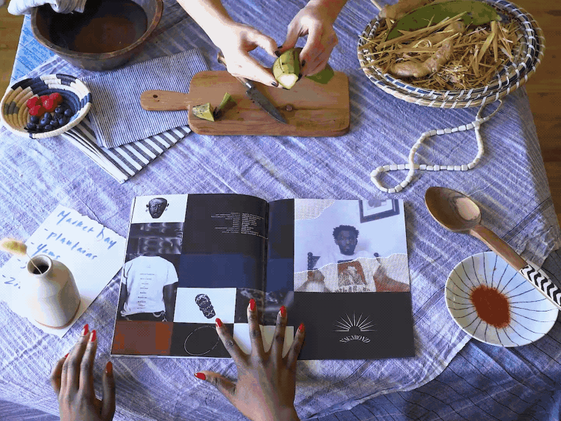 A table with a blue tablecloth featuring various items including a person peeling a banana, a bowl of strawberries and blueberries, a bowl of bananas, a magazine with photos of a person and objects, a note, a small pitcher, a spoon with a striped handle, and a small bowl with a reddish-orange sauce.