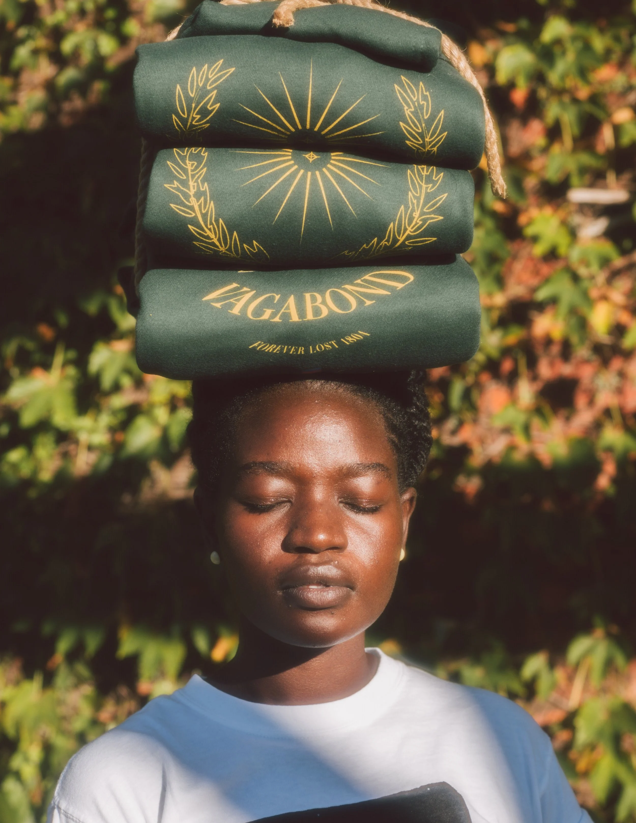 A woman with dark skin and closed eyes balances a stack of green hoodies on her head, with a background of green and brown foliage.