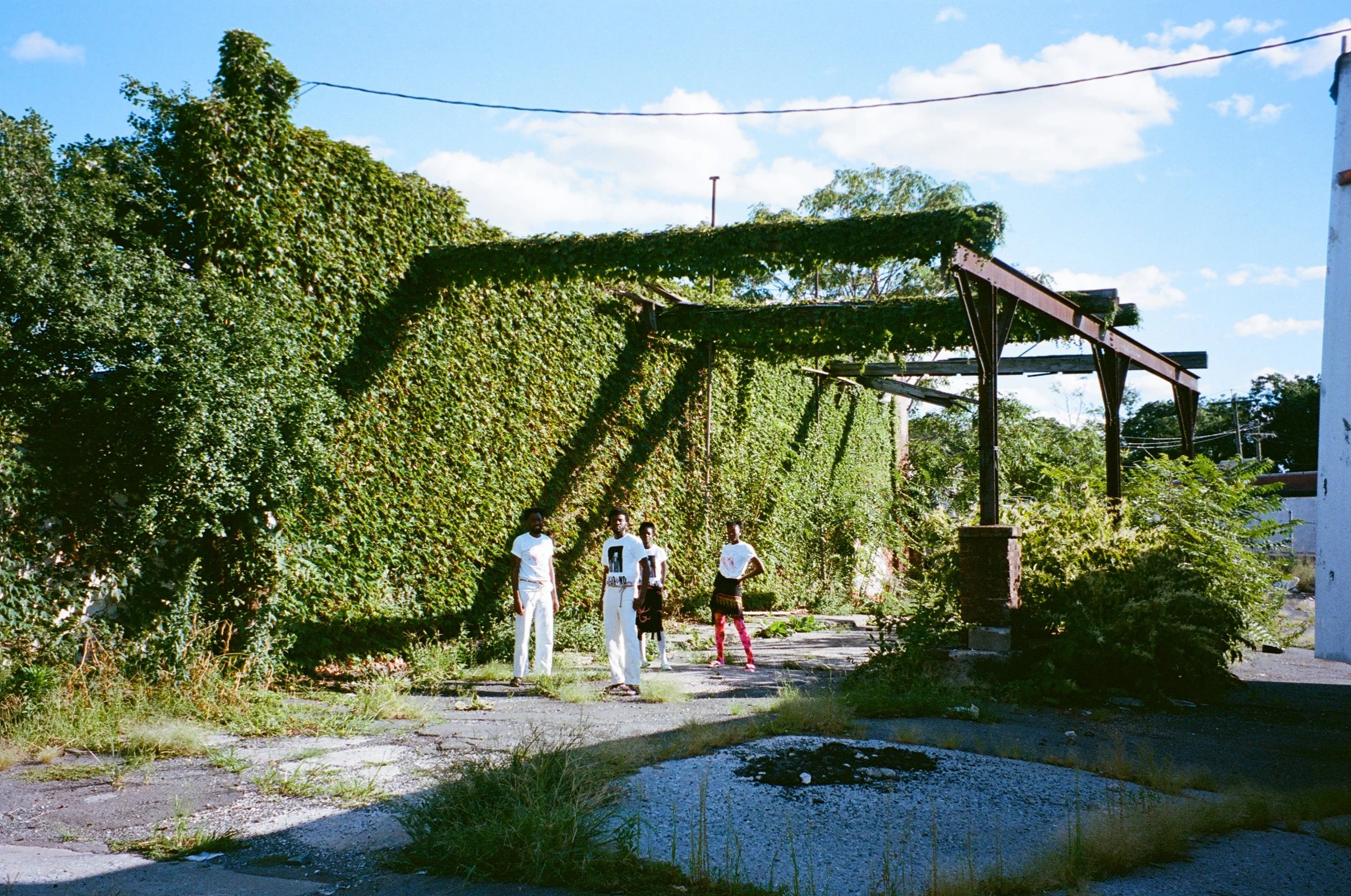Four people standing on an overgrown lot with a tall ivy-covered wall and some metal framework.
