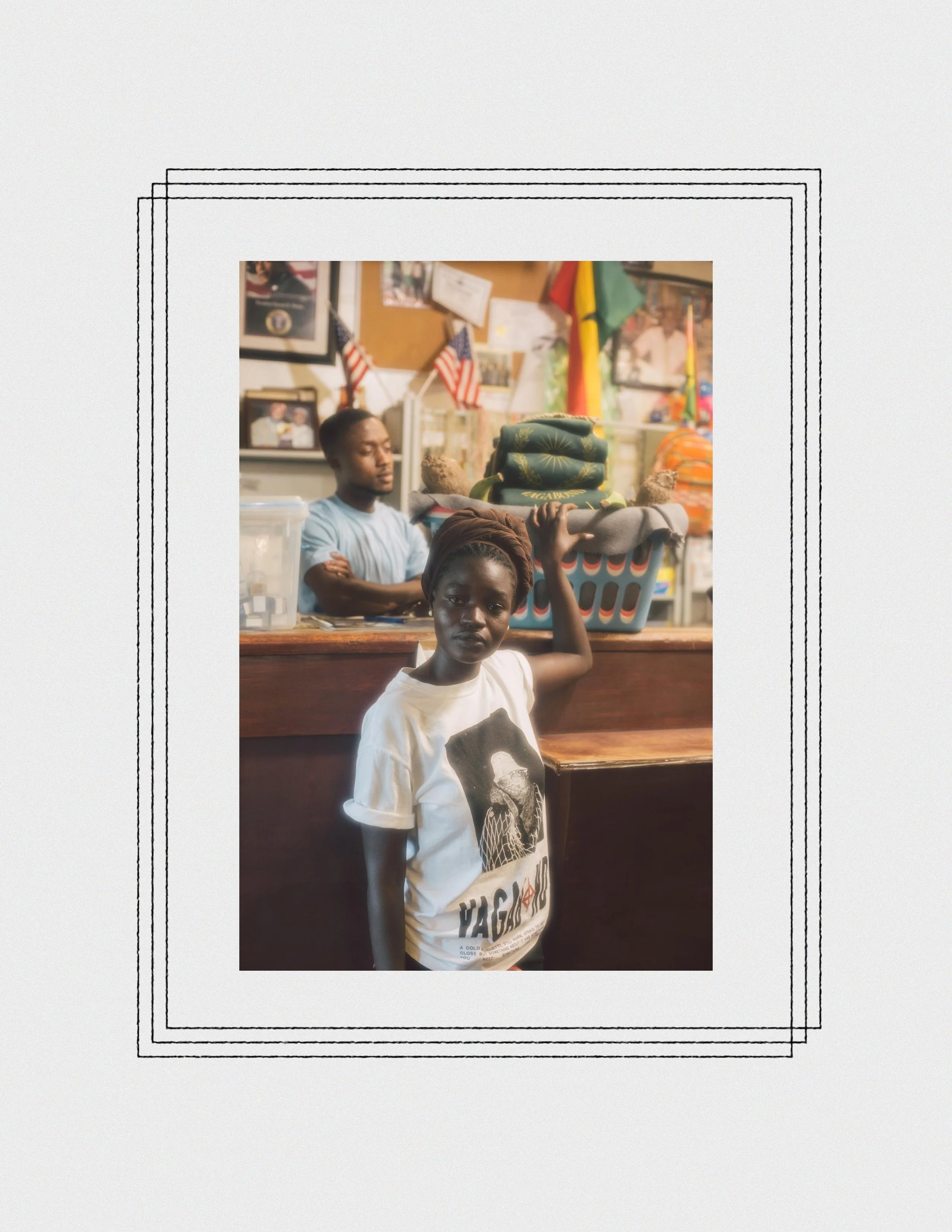 A young girl standing indoors, holding a basket on her head, with a young man in the background. The room has framed pictures and flags on the wall, with colorful decorations.