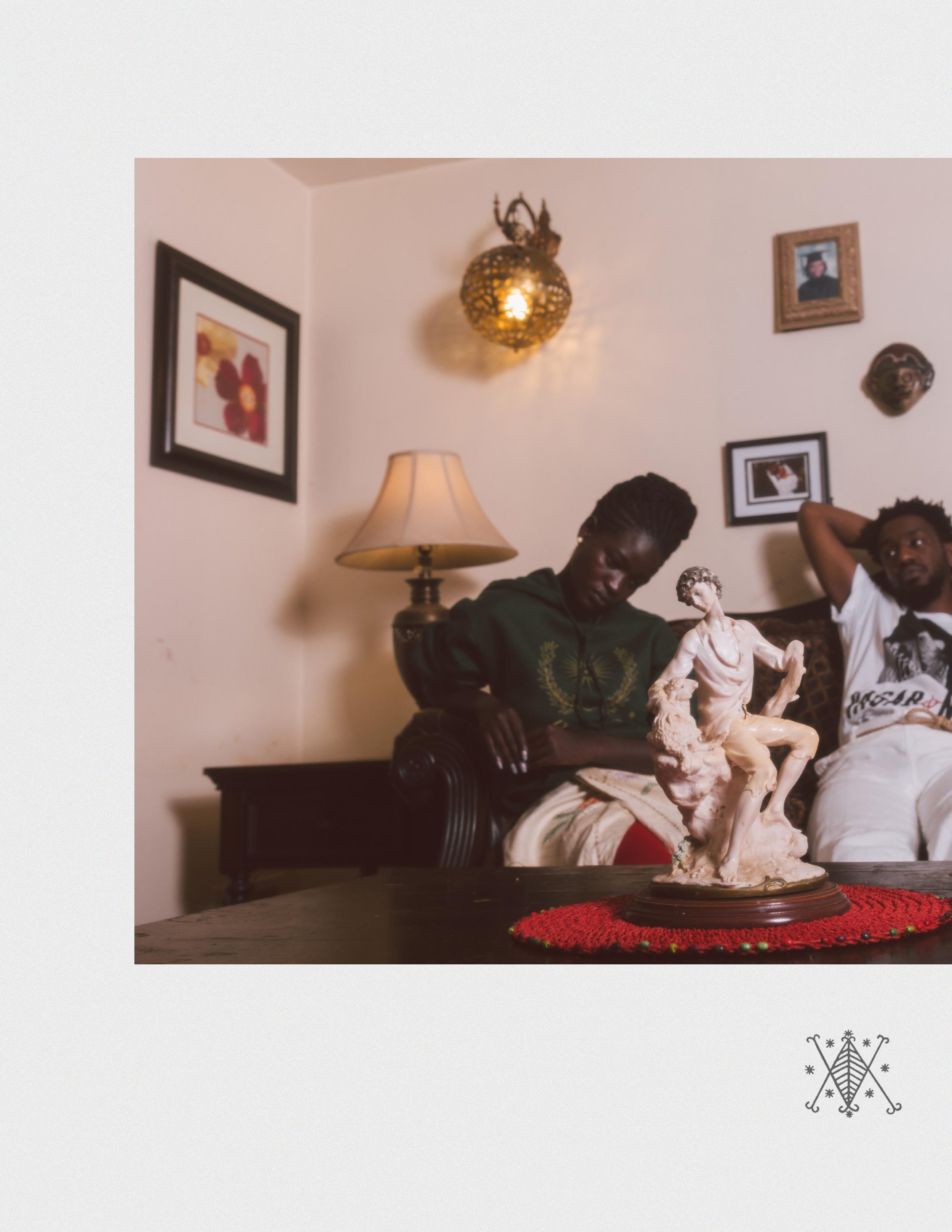 Two people sitting on a couch in a living room with framed pictures and decorative wall lights. A sculpture of a seated man is on a table in front of them.