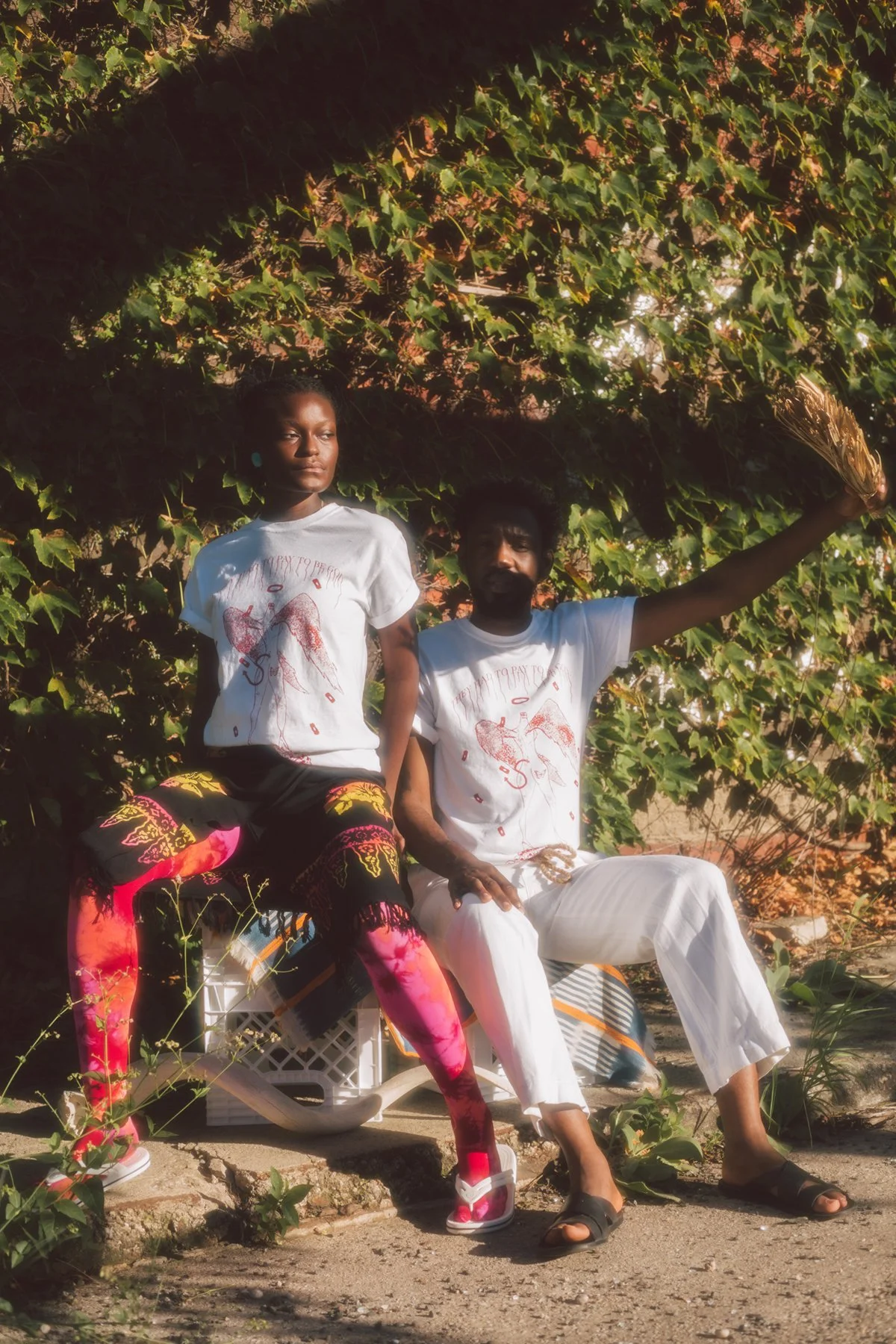 A man and a woman sitting outdoors in front of a leafy hedge. The woman is sitting on a plastic crate, wearing a white t-shirt and colorful pants. The man is seated on the ground, wearing white pants and a white t-shirt, holding a bundle of dried herbs in his right hand and leaning back against the hedge.